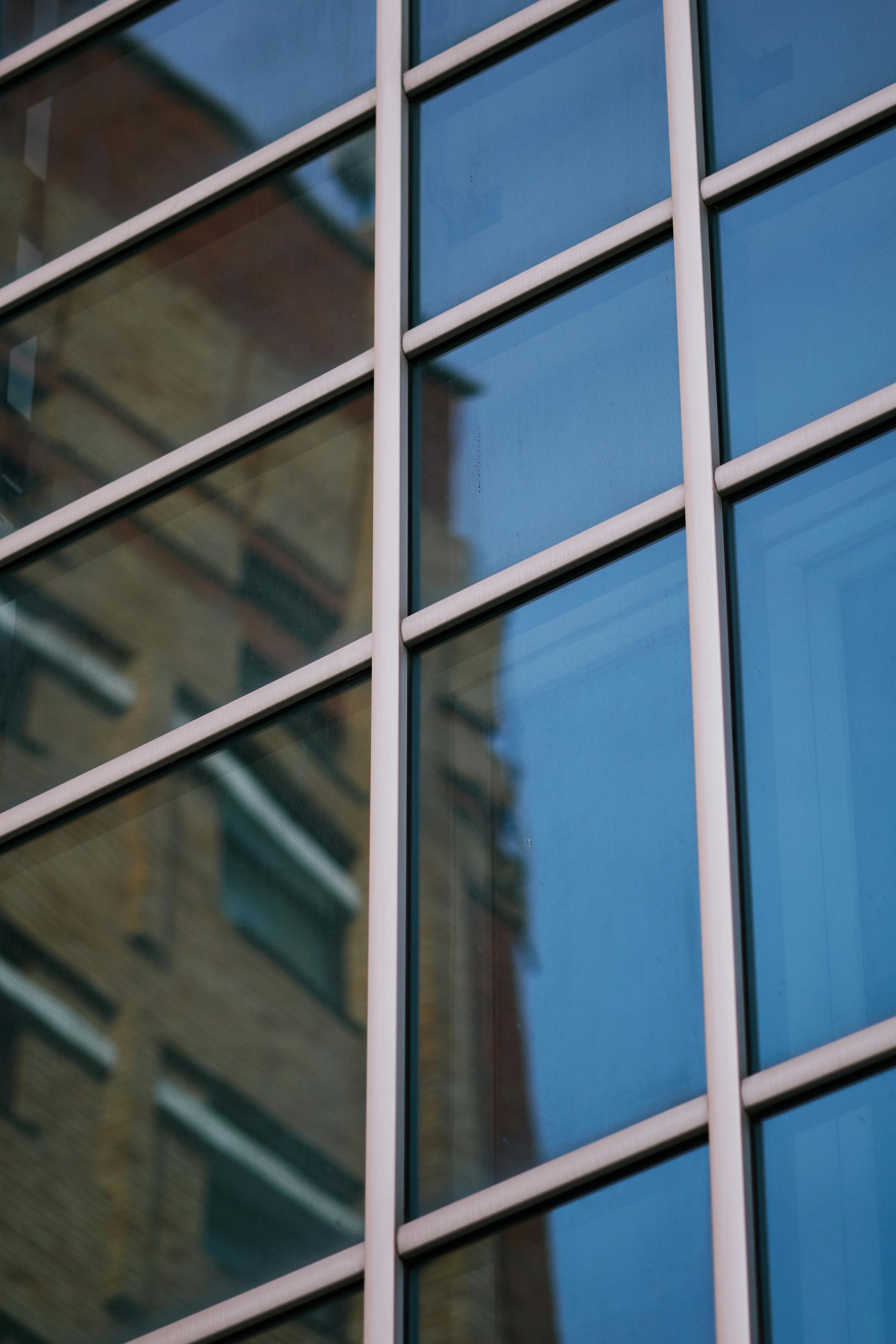 A close-up view of a glass curtain wall with a grid of metal frames reflecting a neighboring brick building.