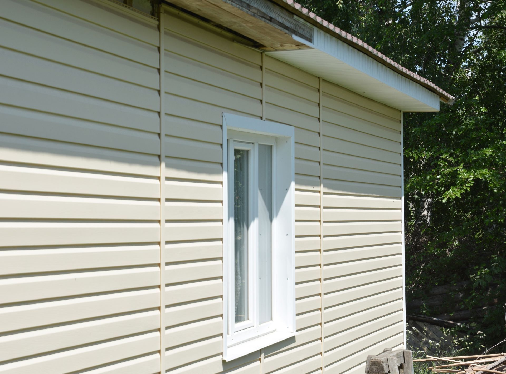 Beige horizontal vinyl siding on an exterior wall with a white window frame under a roof overhang.
