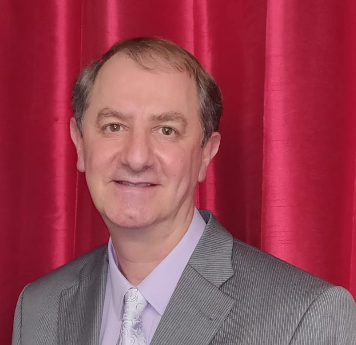 Man in gray suit smiles, standing against a red curtain background.