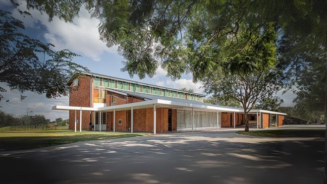 Brick building with a flat roof and long covered walkway; trees frame the structure against a blue sky.