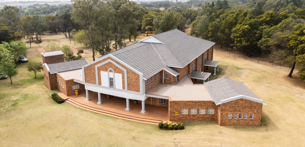 An aerial view of a brick building with a gray roof, surrounded by green trees and grass.