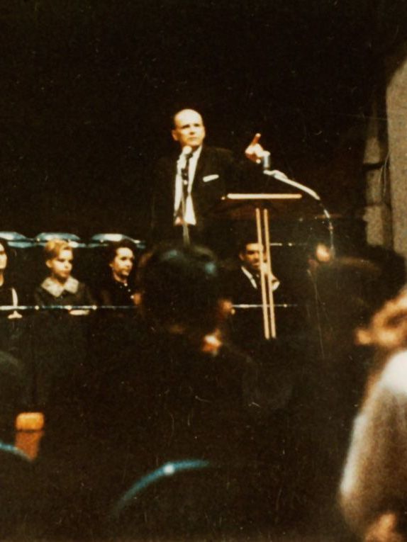 Man speaking at a podium, gesturing. Audience members seated in front. Dim setting.