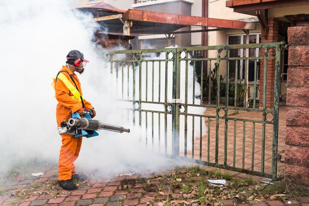 A Man Is Spraying Smoke On A Fence In Front Of A House — ASAP Pest Control in Booval, QLD