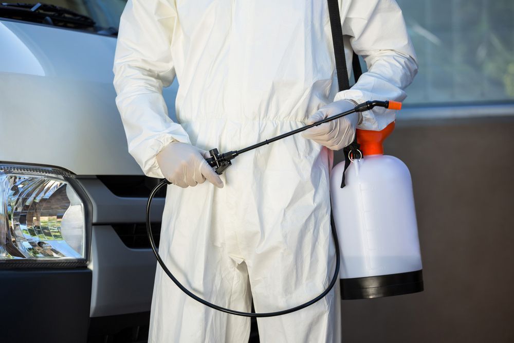 A Man In A Protective Suit Is Holding A Sprayer In Front Of A Van — ASAP Pest Control in Logan, QLD