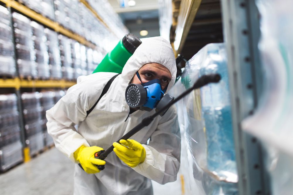 A Man In A Protective Suit And Mask Is Spraying Chemicals In A Warehouse — ASAP Pest Control in Elanora, QLD