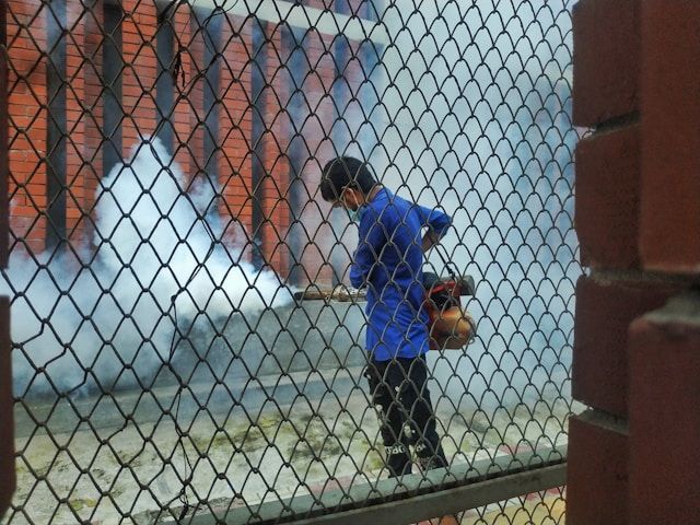 A Man In A Blue Shirt Is Spraying Smoke Through A Chain Link Fence — ASAP Pest Control in Rathdowney, QLD