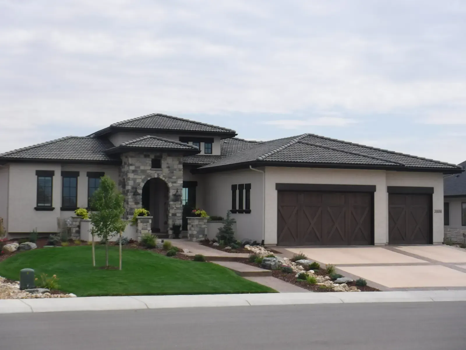 Beige stucco house with stone accents, brown garage doors, and a tile roof on a cloudy day.