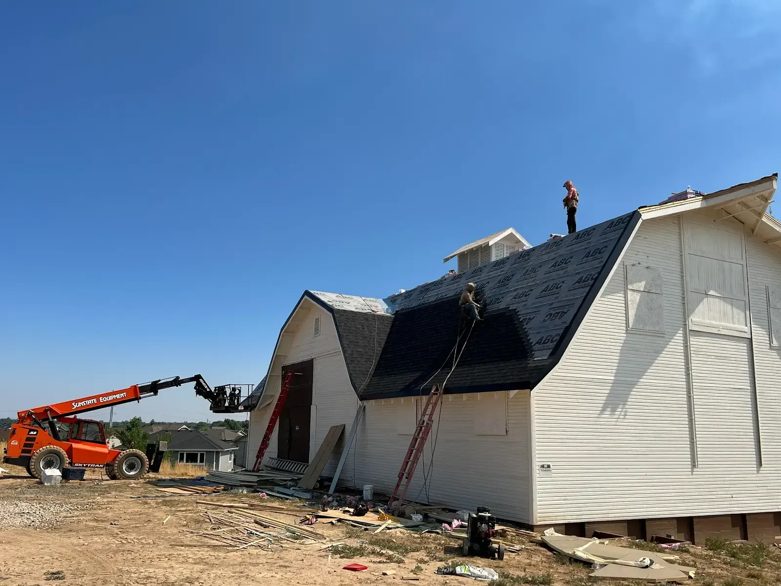 Construction of a white barn with a dark roof. A person works on the roof with a lift nearby.