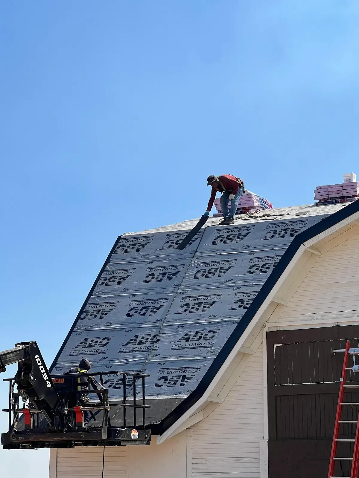 Workers installing roofing on a white barn under a blue sky, one in a lift.