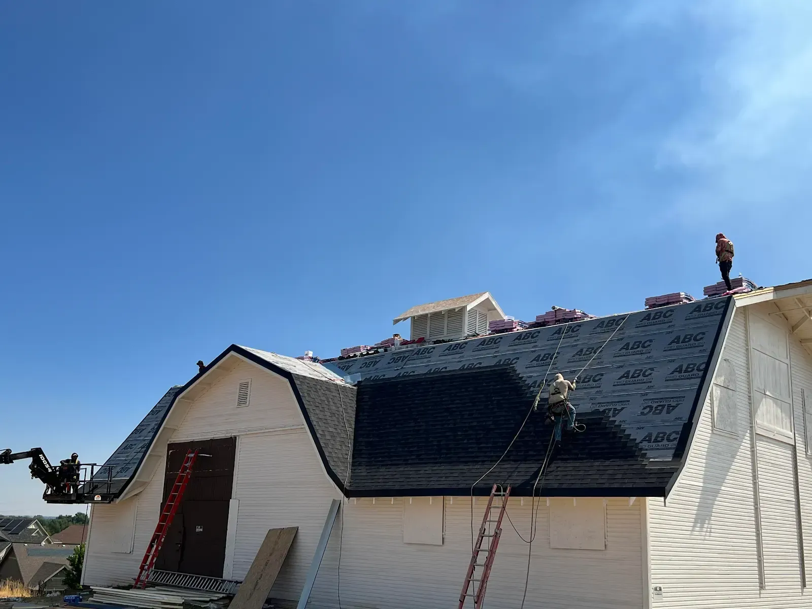 Workers on a roof installing shingles on a white building against a clear blue sky.