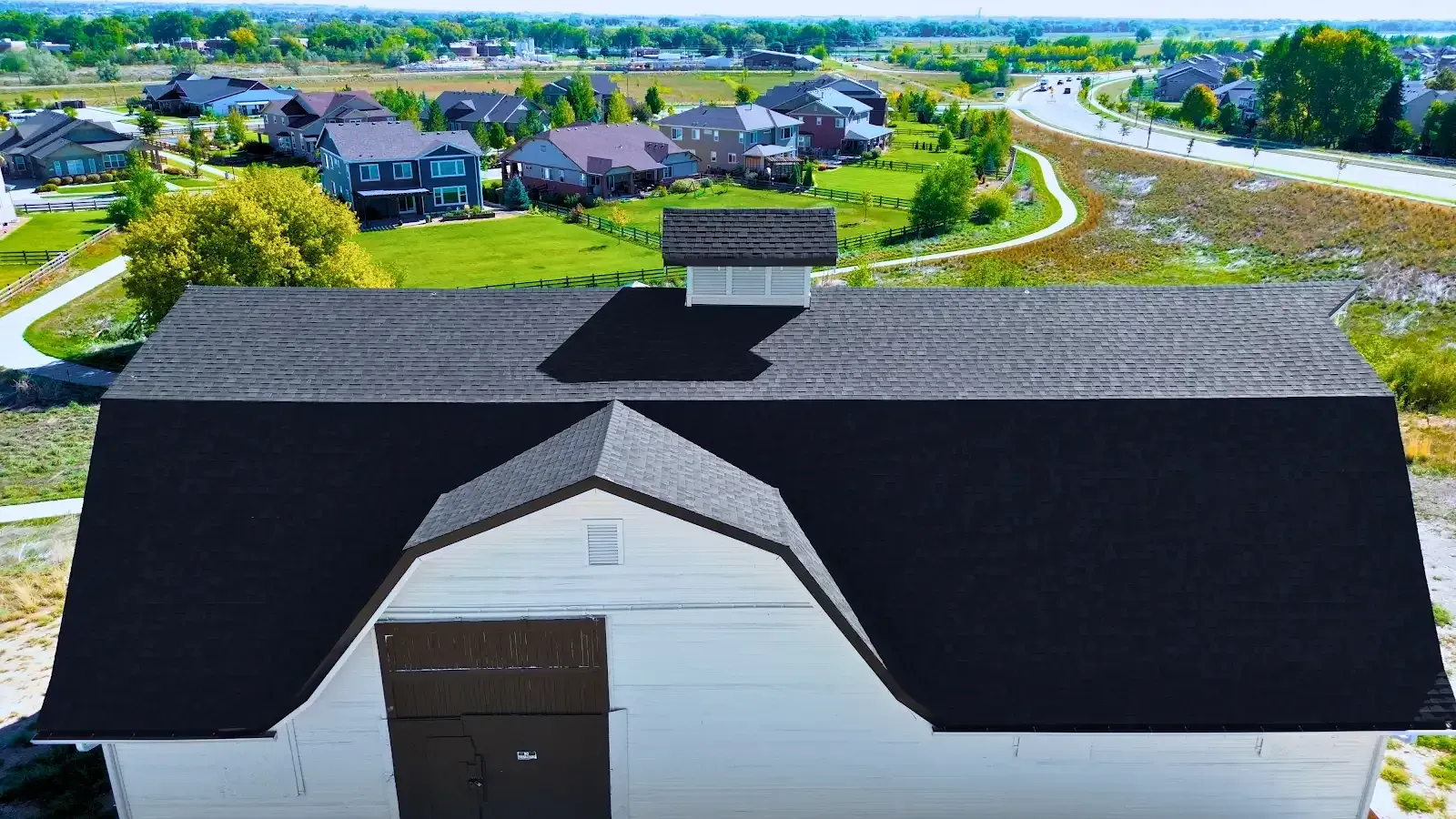 White barn with black roof, small cupola, against a backdrop of houses and green landscape.
