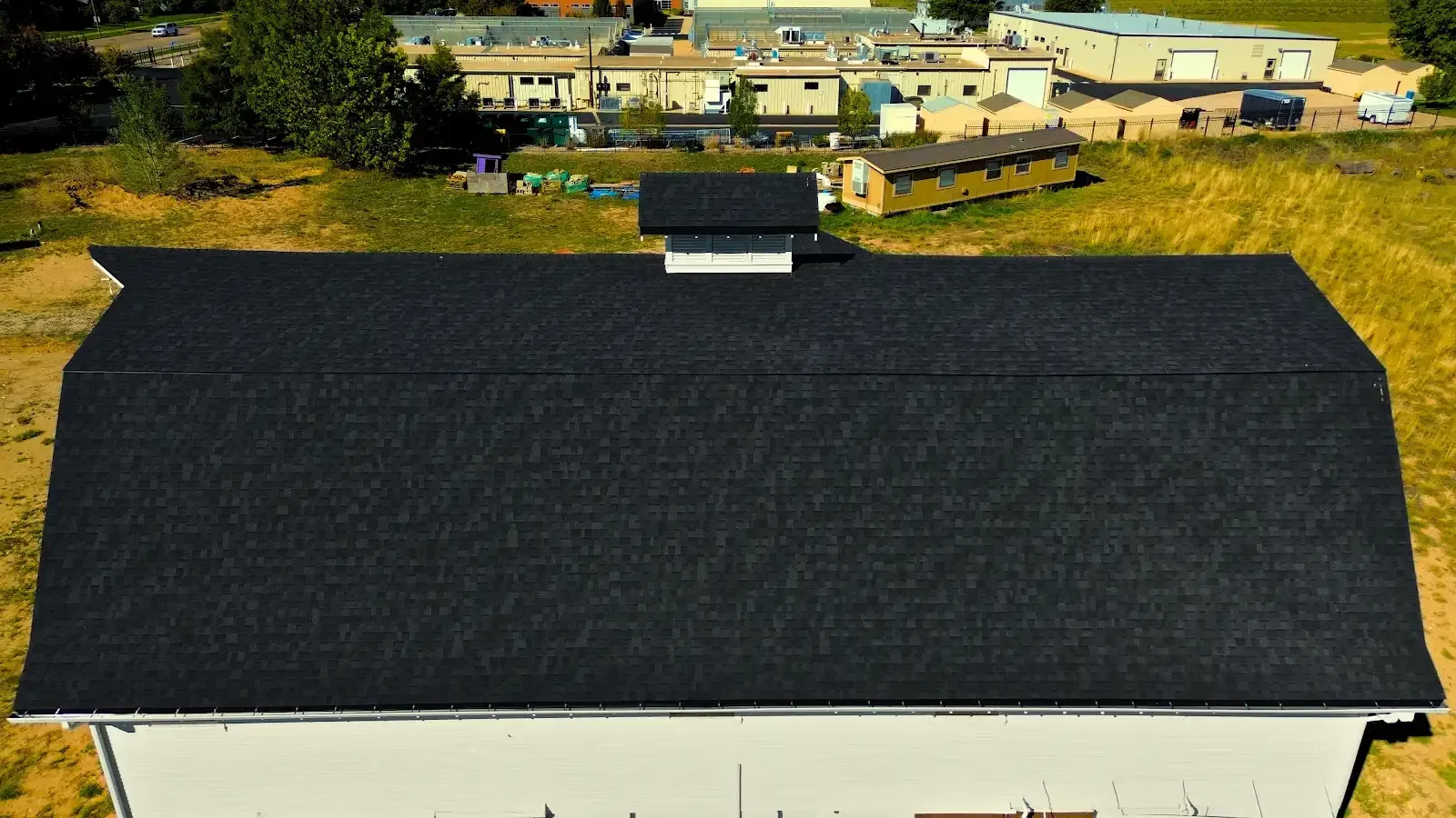 Black roof of a white barn, overhead view. Green field and buildings in background.