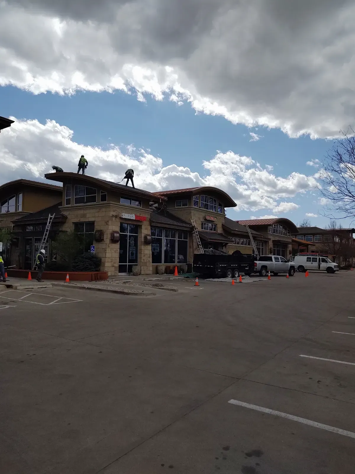 Workers on a stone building roof under a cloudy sky. Cars and cones are in front.