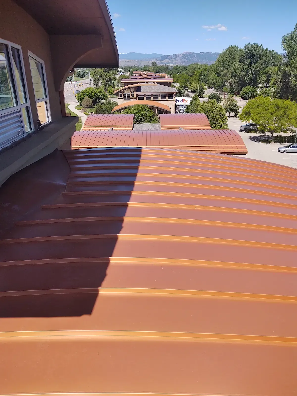 A copper-colored corrugated roof on a building with a view of other buildings and trees on a sunny day.