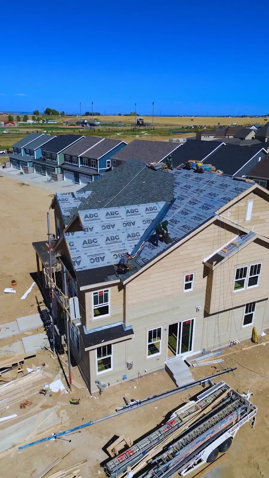 Construction of a two-story beige house; roofers working on the rooftop. Other new houses visible in background.