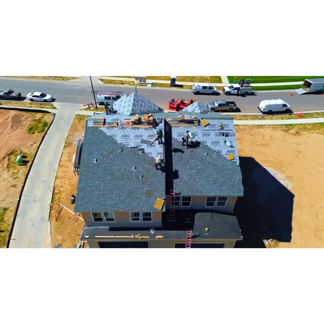 Workers installing roof shingles on a two-story house, with construction materials and vehicles nearby.