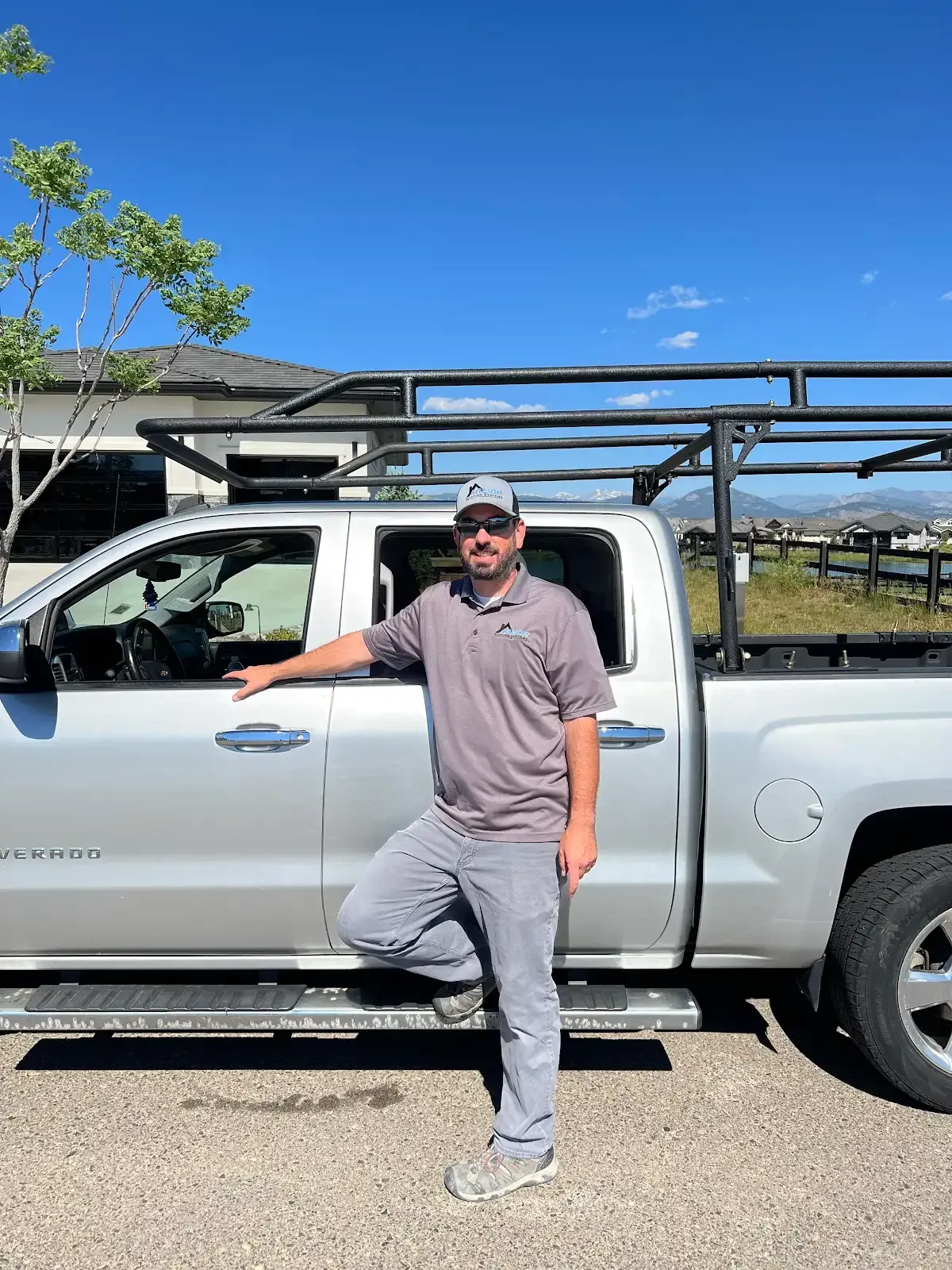 Man in gray work clothes stands next to silver pickup truck with a ladder rack, under a blue sky.