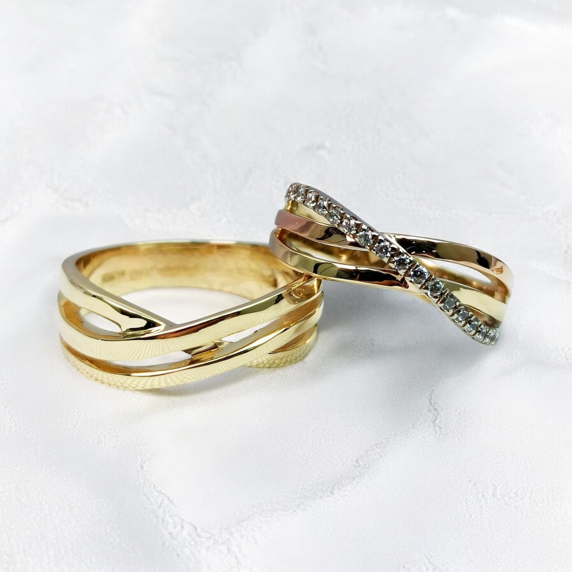 Dos anillos cruzados de oro, uno con cristales, sobre una superficie texturizada blanca.