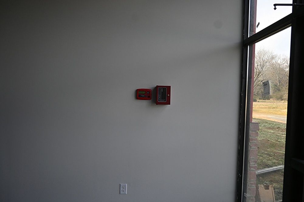 A red fire alarm is hanging on a white wall next to a window.