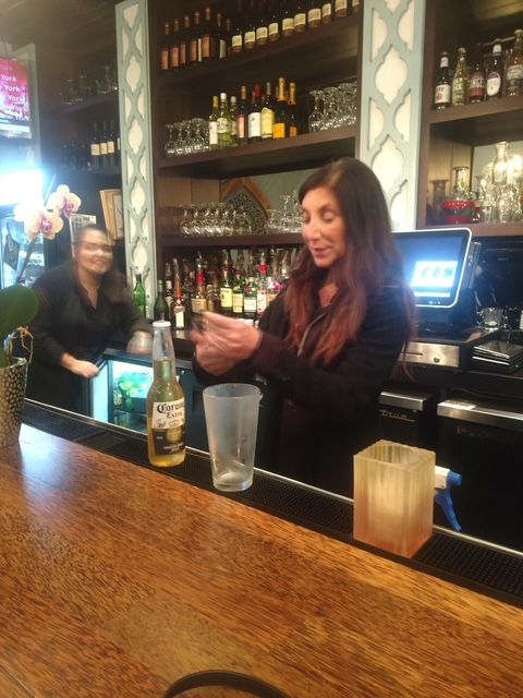A woman is standing behind a bar holding a bottle of corona
