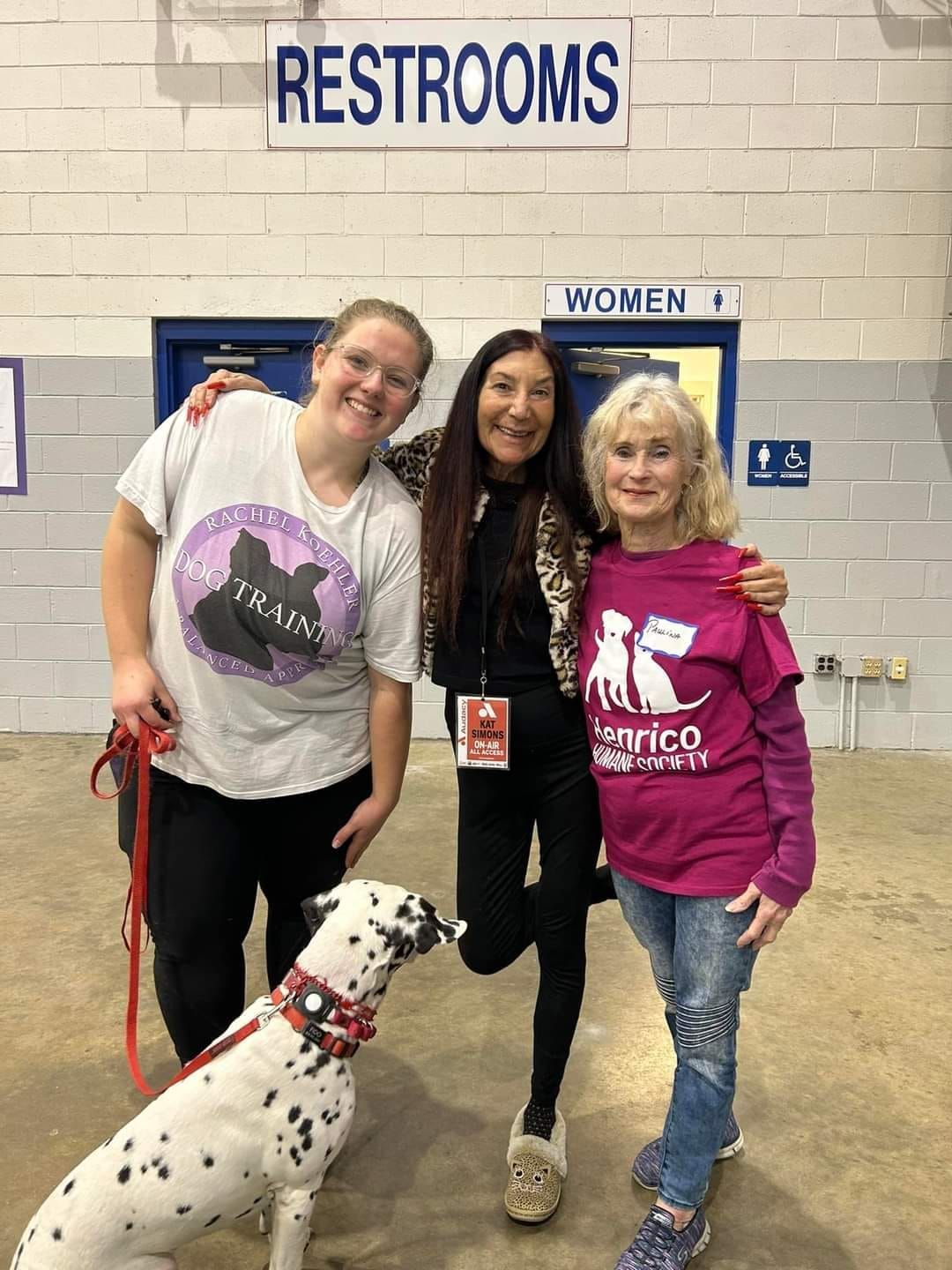 Three women are posing for a picture with a dalmatian dog in front of a restrooms sign.