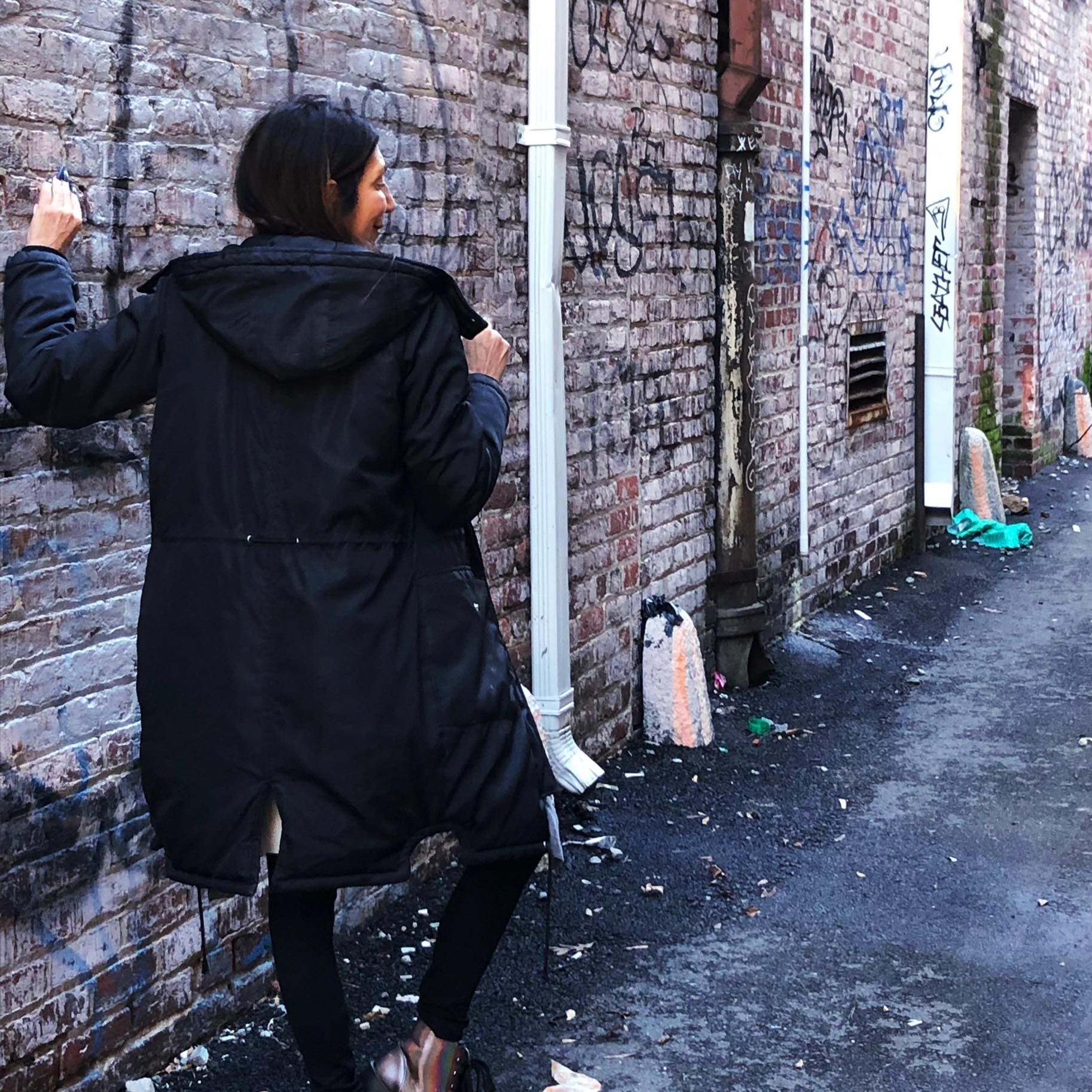 A woman in a black coat is leaning against a brick wall