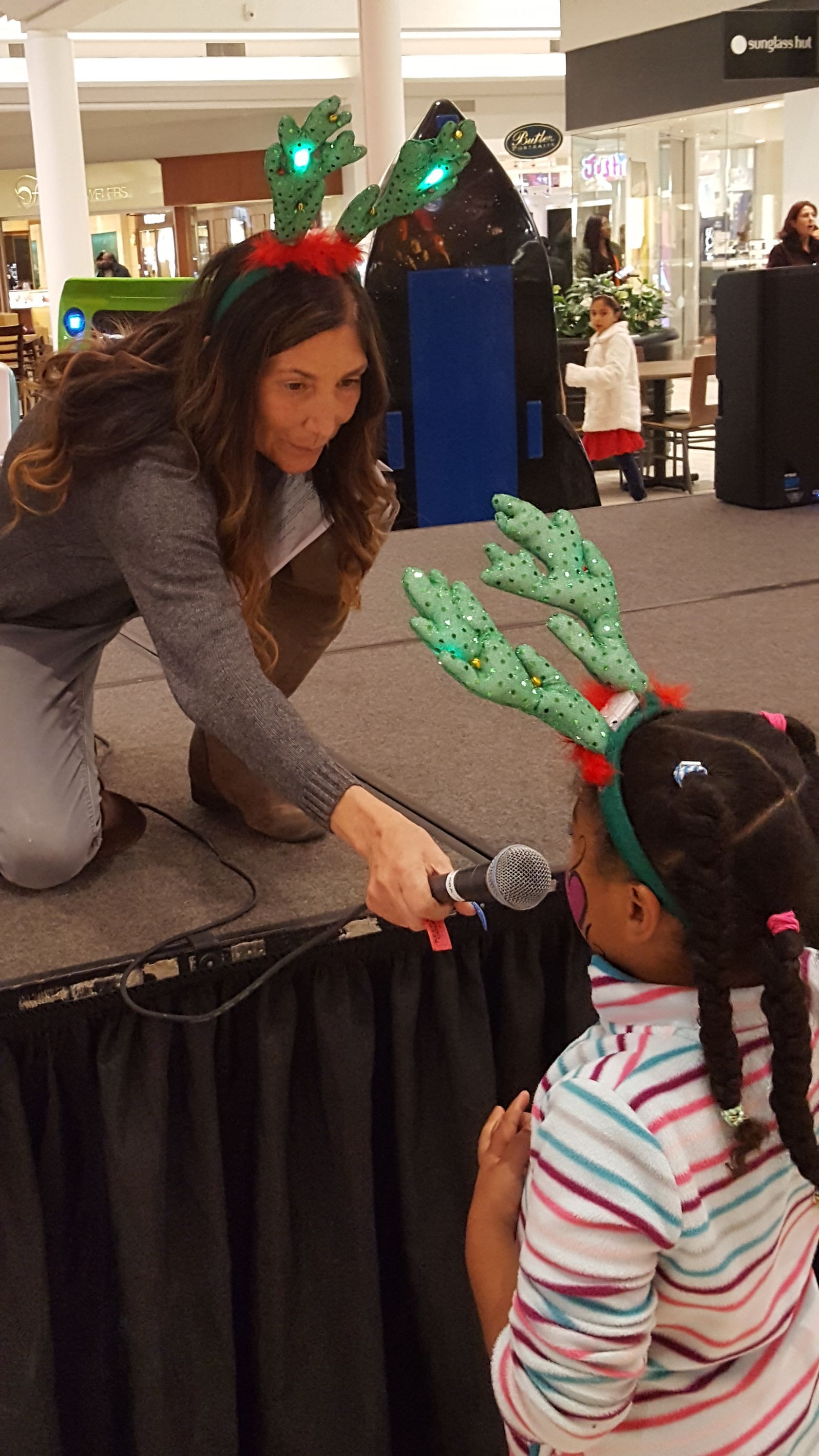 A woman is kneeling down next to a little girl wearing a reindeer headband.