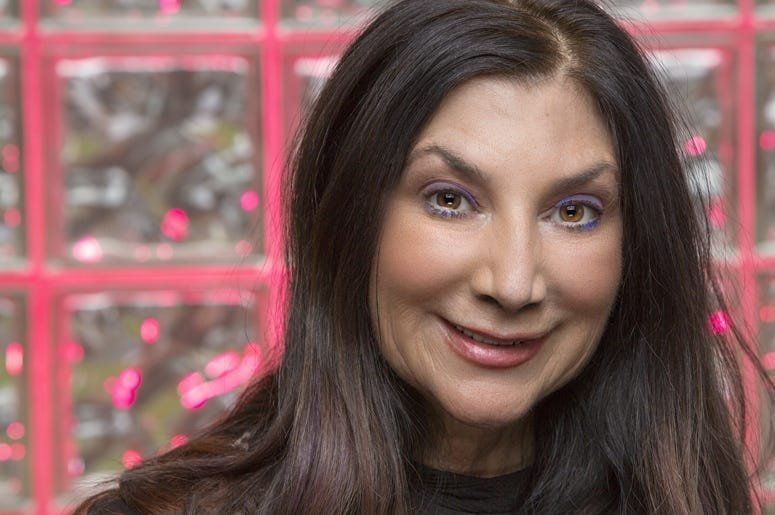 A woman with long dark hair is smiling in front of a pink wall.