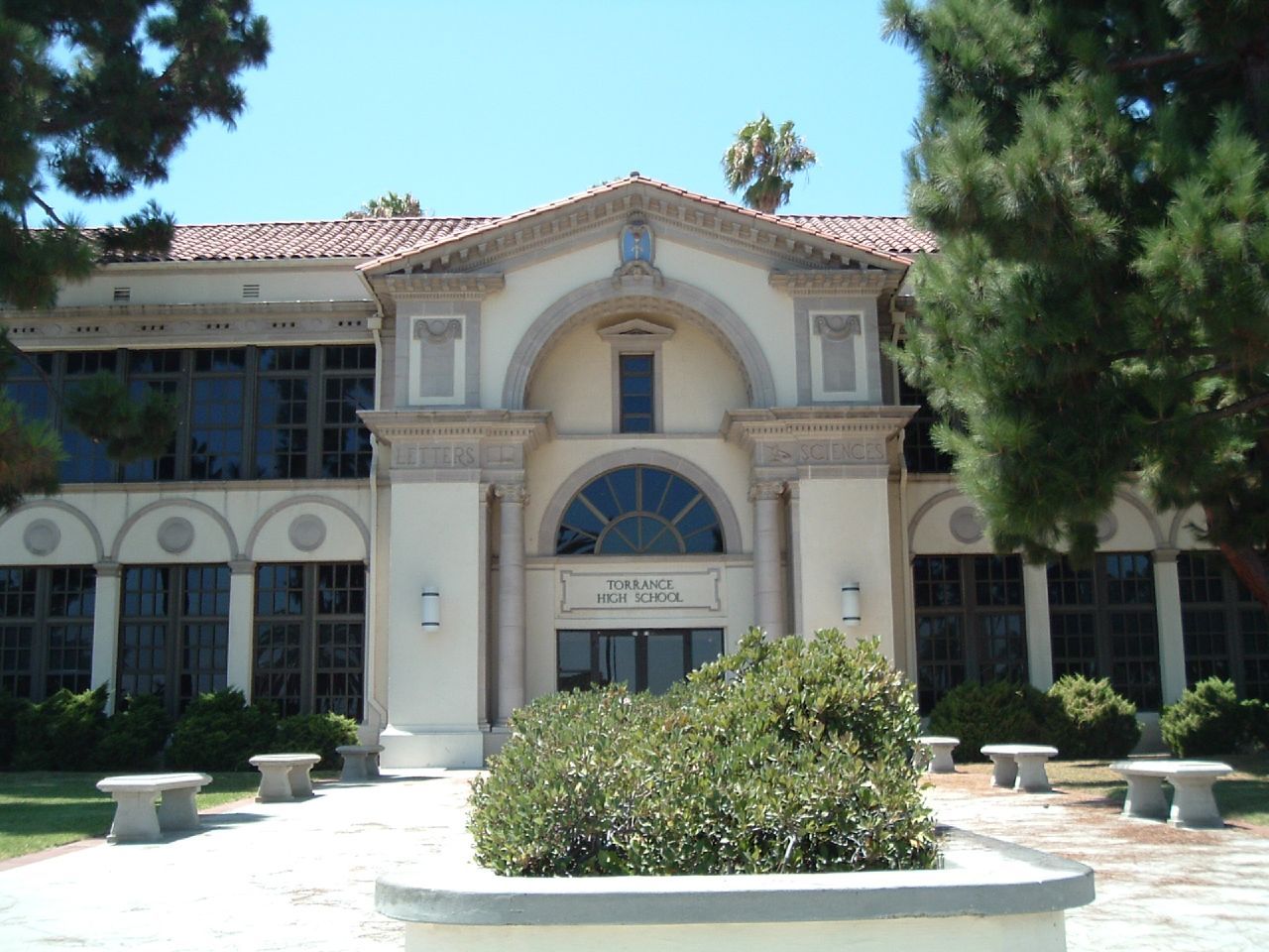 Front entrance of a light-colored building with arched doorway and large windows, surrounded by trees and a sunny sky.
