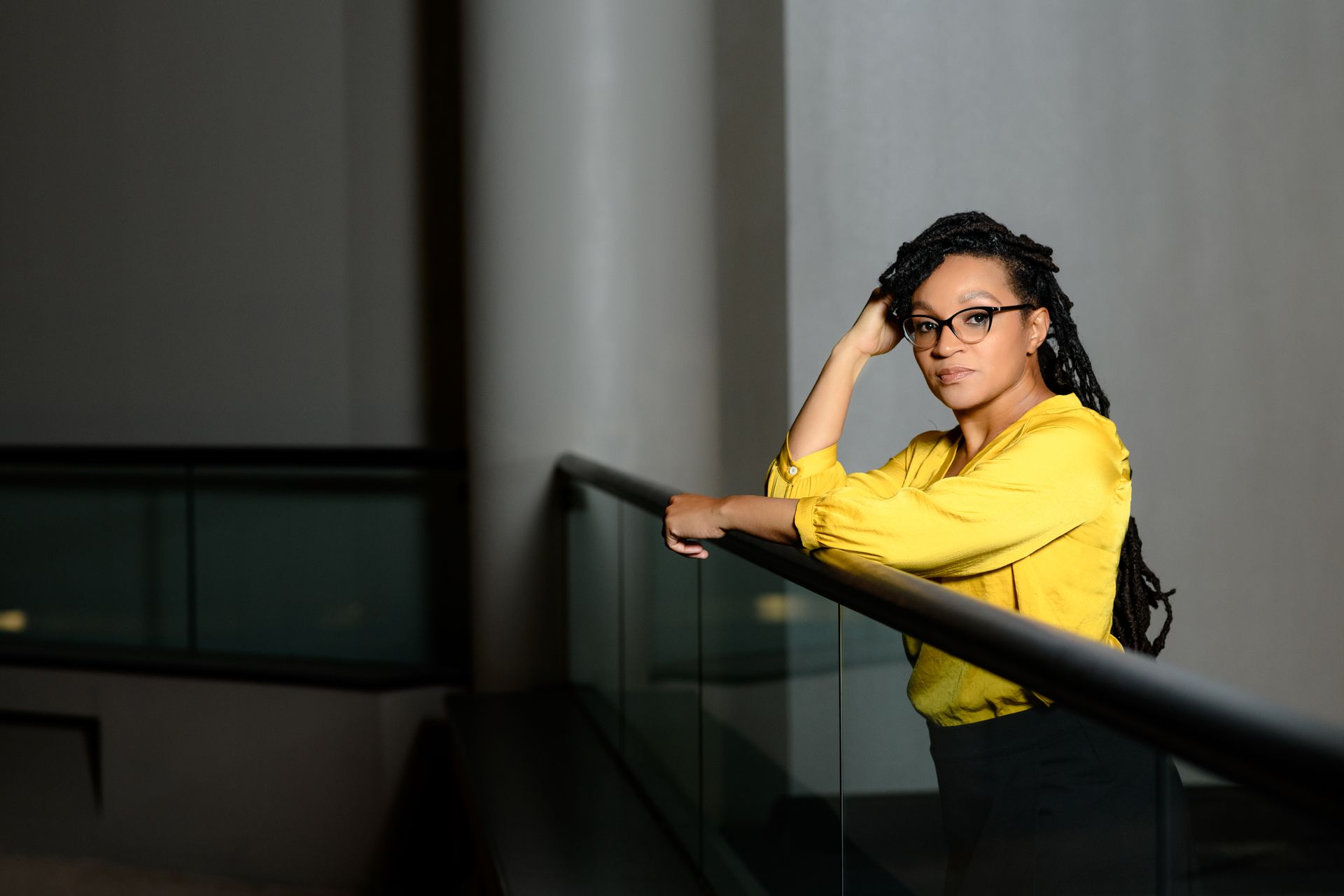 A woman in a yellow shirt and glasses is leaning on a railing.