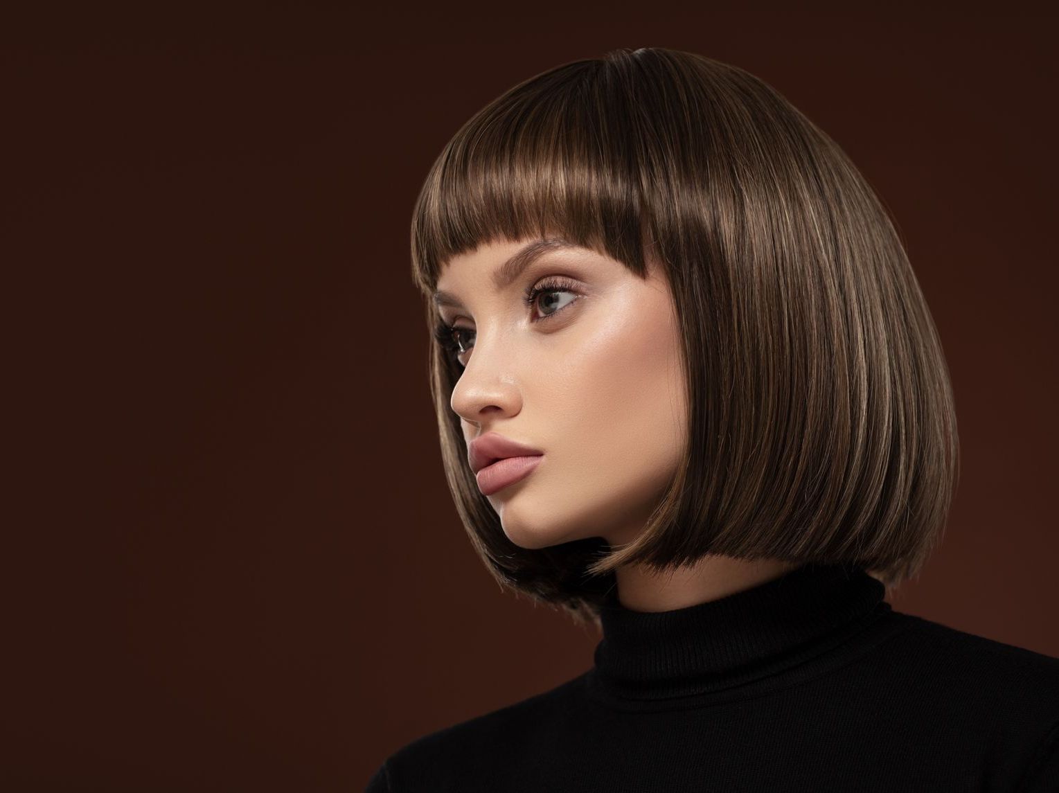 Woman with short, brown bob haircut, blunt bangs, wearing a black turtleneck against a brown background.