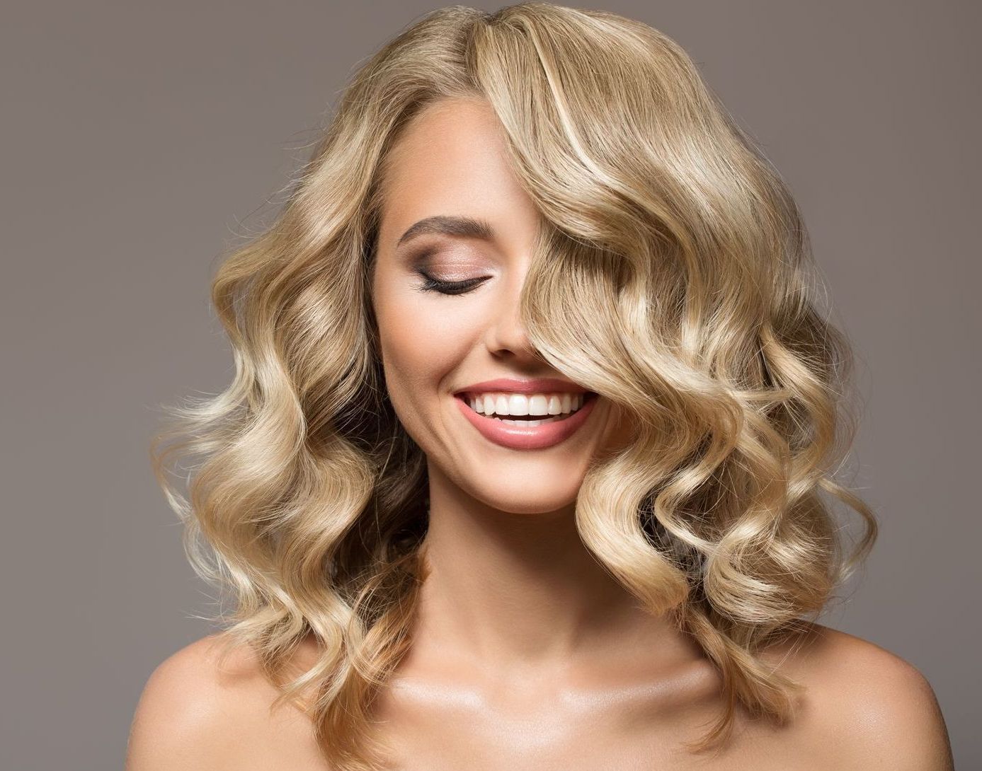 Blonde woman with wavy hair smiling, eyes closed, against a neutral background.