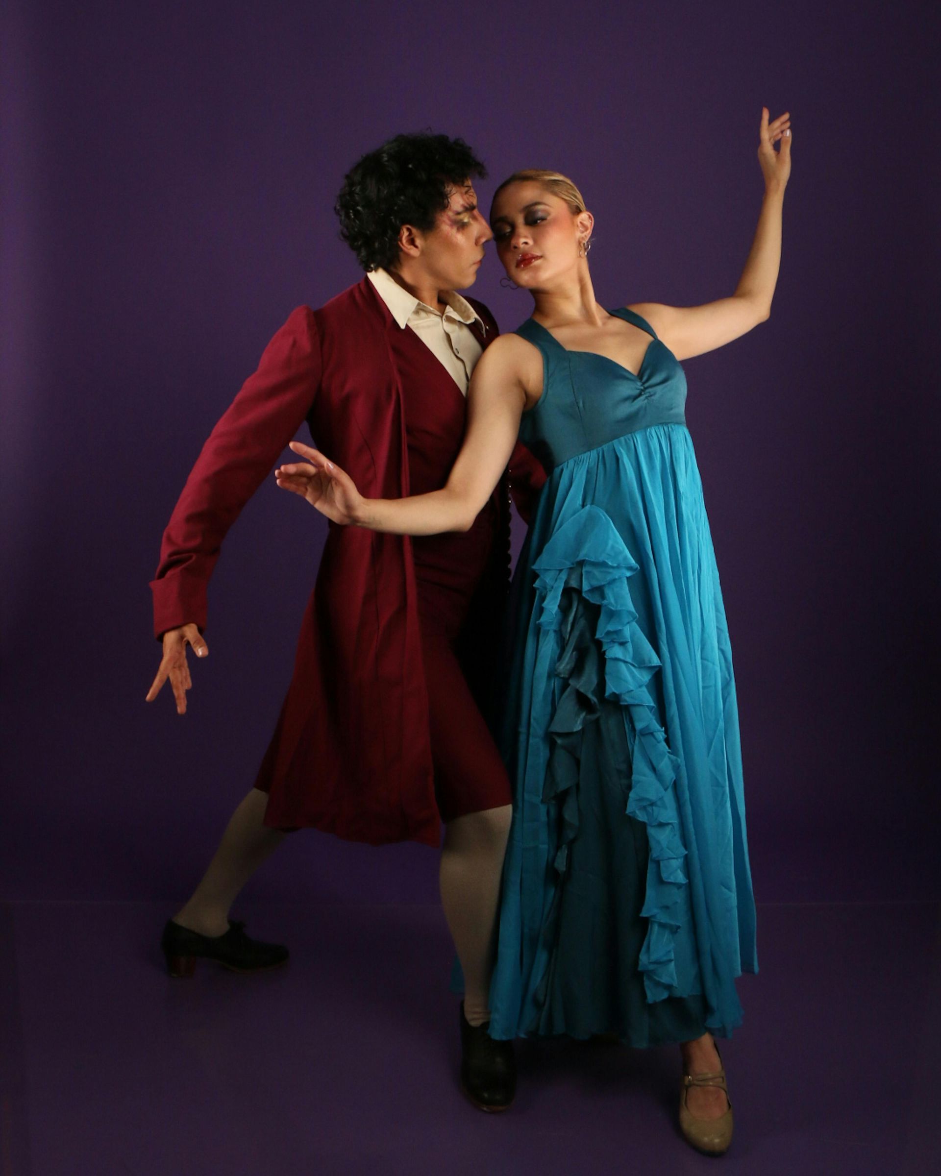 Two dancers in performance. Man in red coat, woman in blue dress. Purple background.