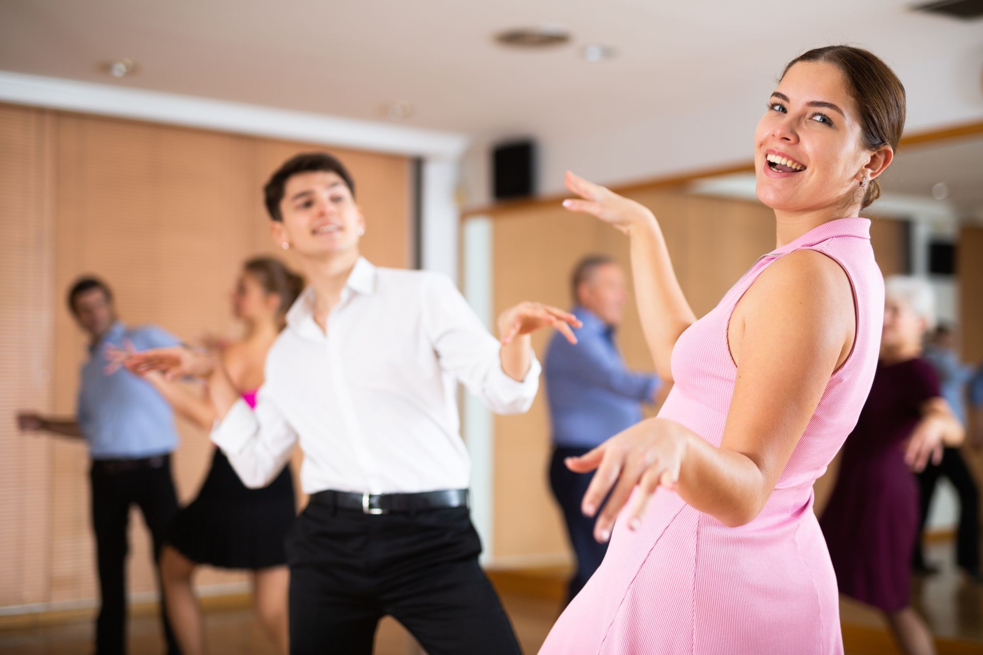 People dancing in a dance studio, with a smiling woman in pink leading.