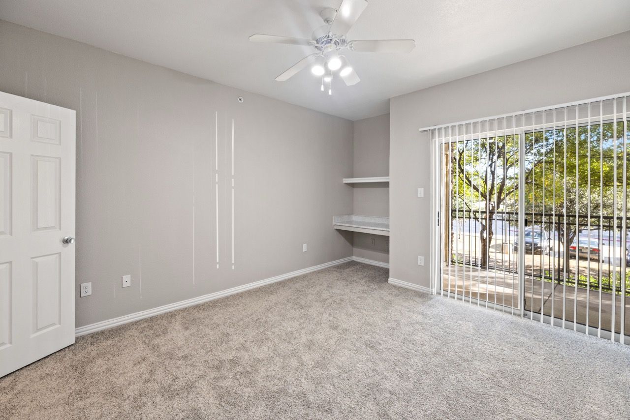 Empty apartment room with carpet, ceiling fan, and sliding glass door with vertical blinds.