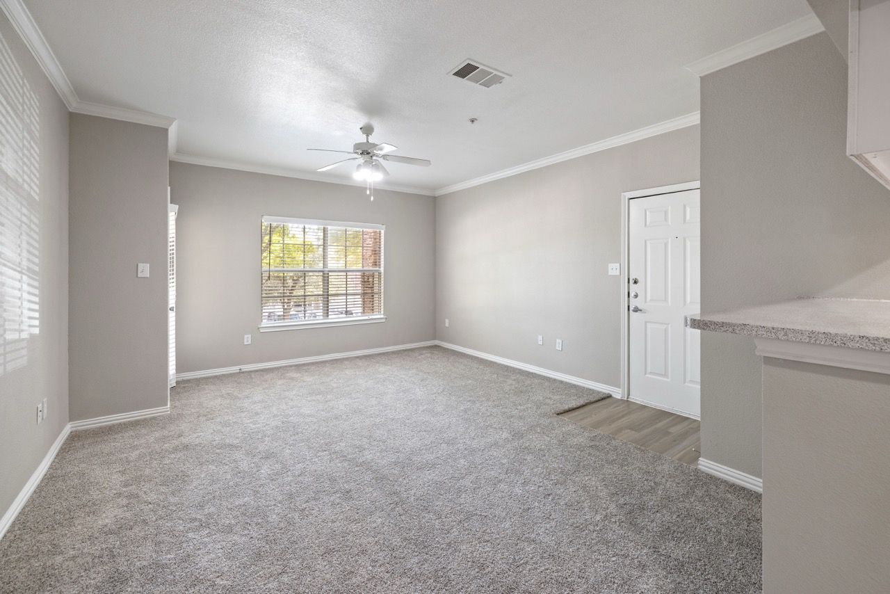 Neutral living room with carpet, window blinds, a ceiling fan, and an open kitchen counter.