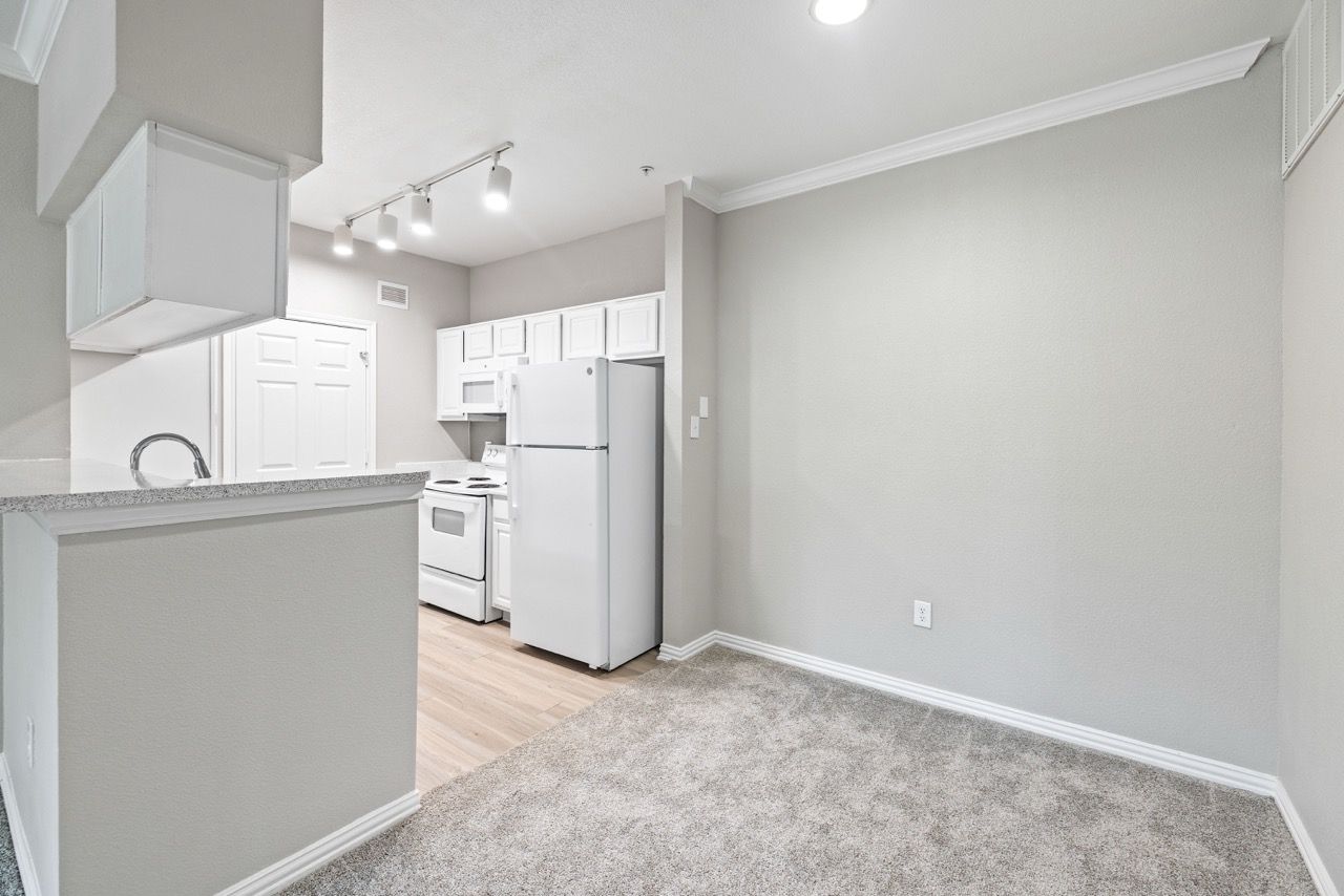 Interior of a modern apartment kitchen with white cabinets and a fridge, adjacent carpeted living area.