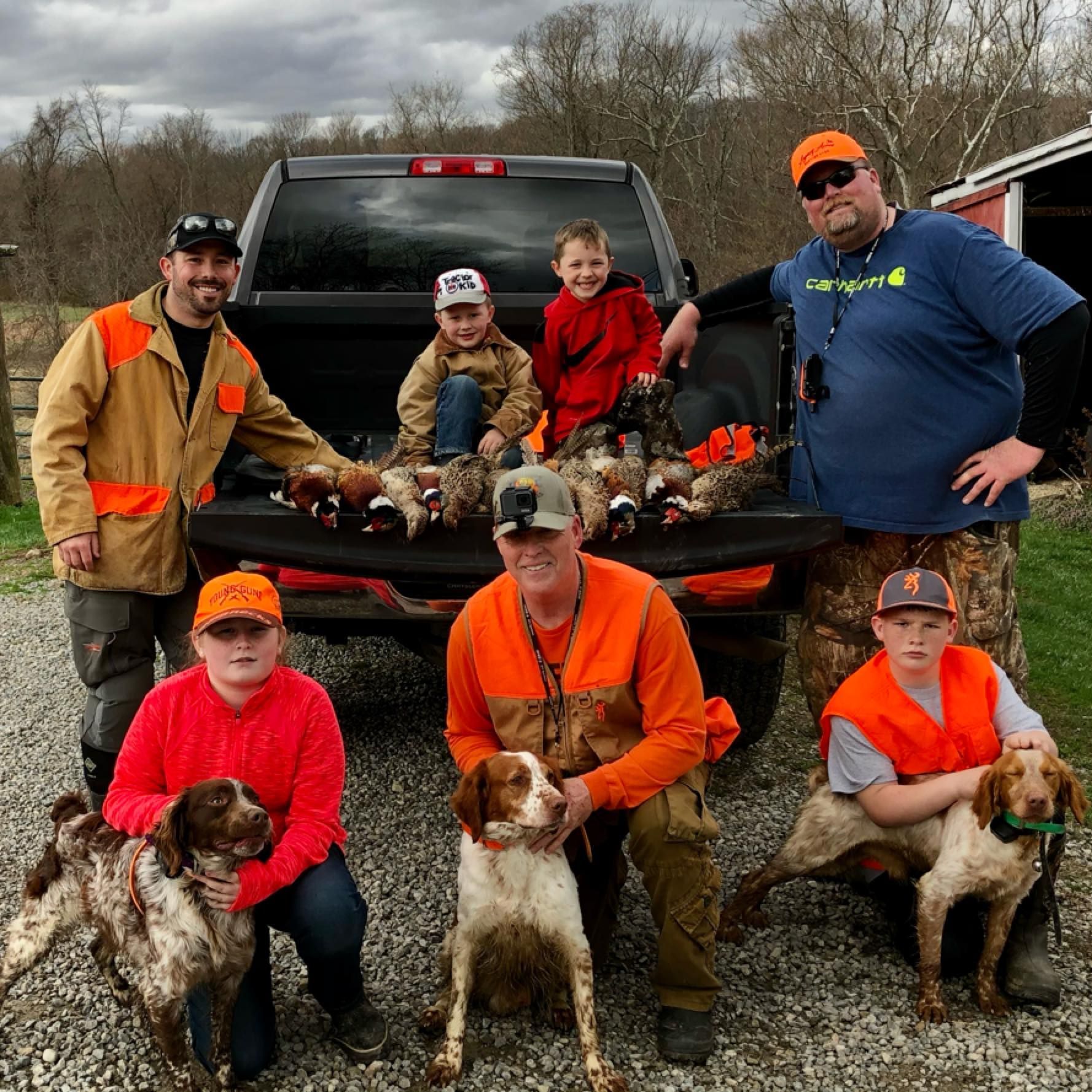Group of people and dogs with hunted birds in a truck bed, all wearing safety orange, in a rural setting.