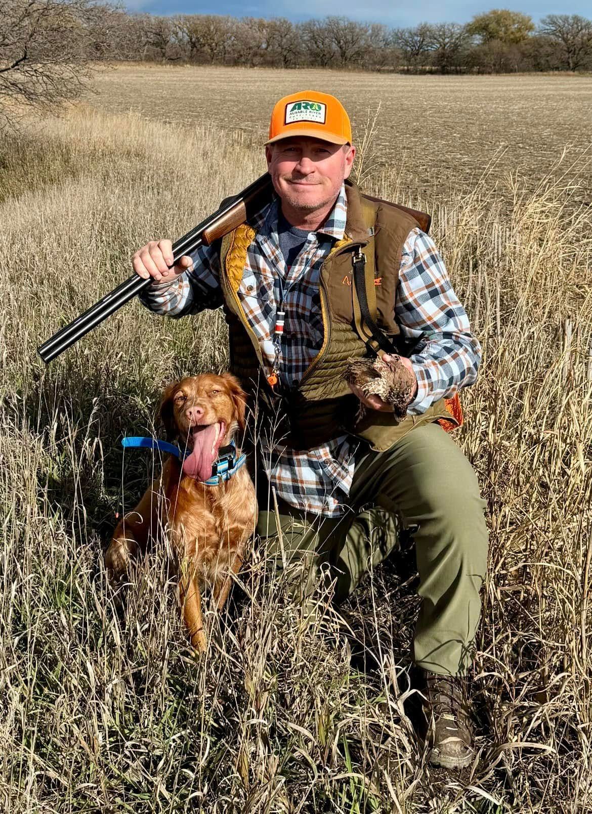 Man kneeling in field with a dog, holding a shotgun and a bird. Orange hat and vest, fall setting.
