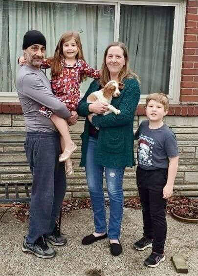 Family of four poses outdoors: man holding a child, woman holding a dog, and a child standing.