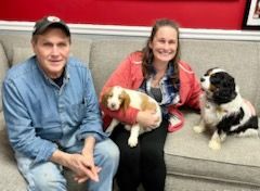 Man and woman on a couch with two Cavalier King Charles Spaniel puppies. Woman holds one puppy. All are smiling.