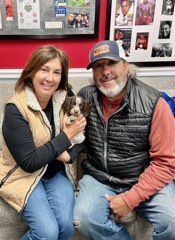 Couple holding a small, brown and white dog. They are smiling. Indoor setting with photos on the wall.