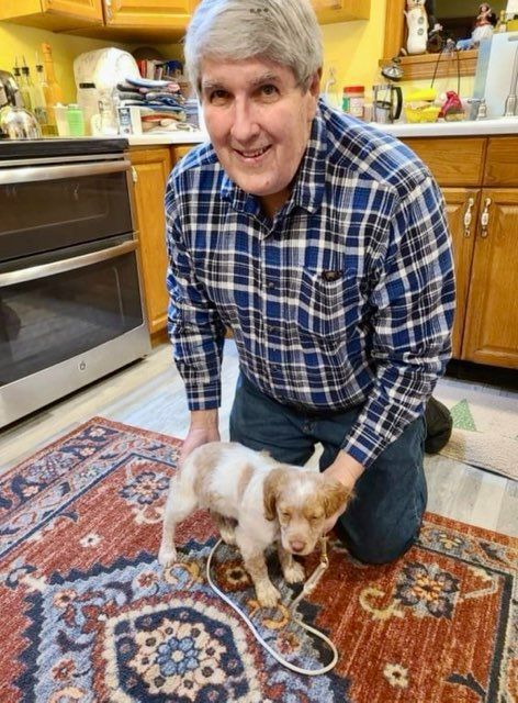 Man kneeling next to puppy on patterned rug in kitchen, smiling.