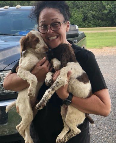 Woman holding two small dogs, smiling broadly. The dogs have brown and white fur, and appear cuddled up.