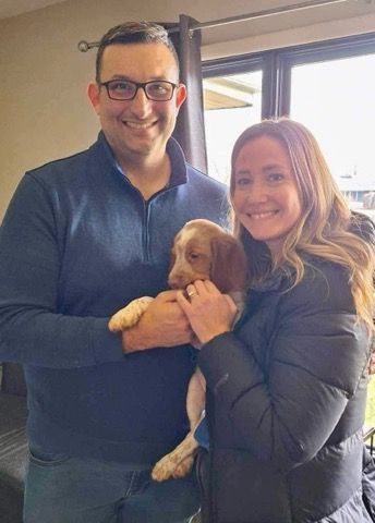 Couple holding a puppy indoors. Man in blue sweater, woman in a black jacket.