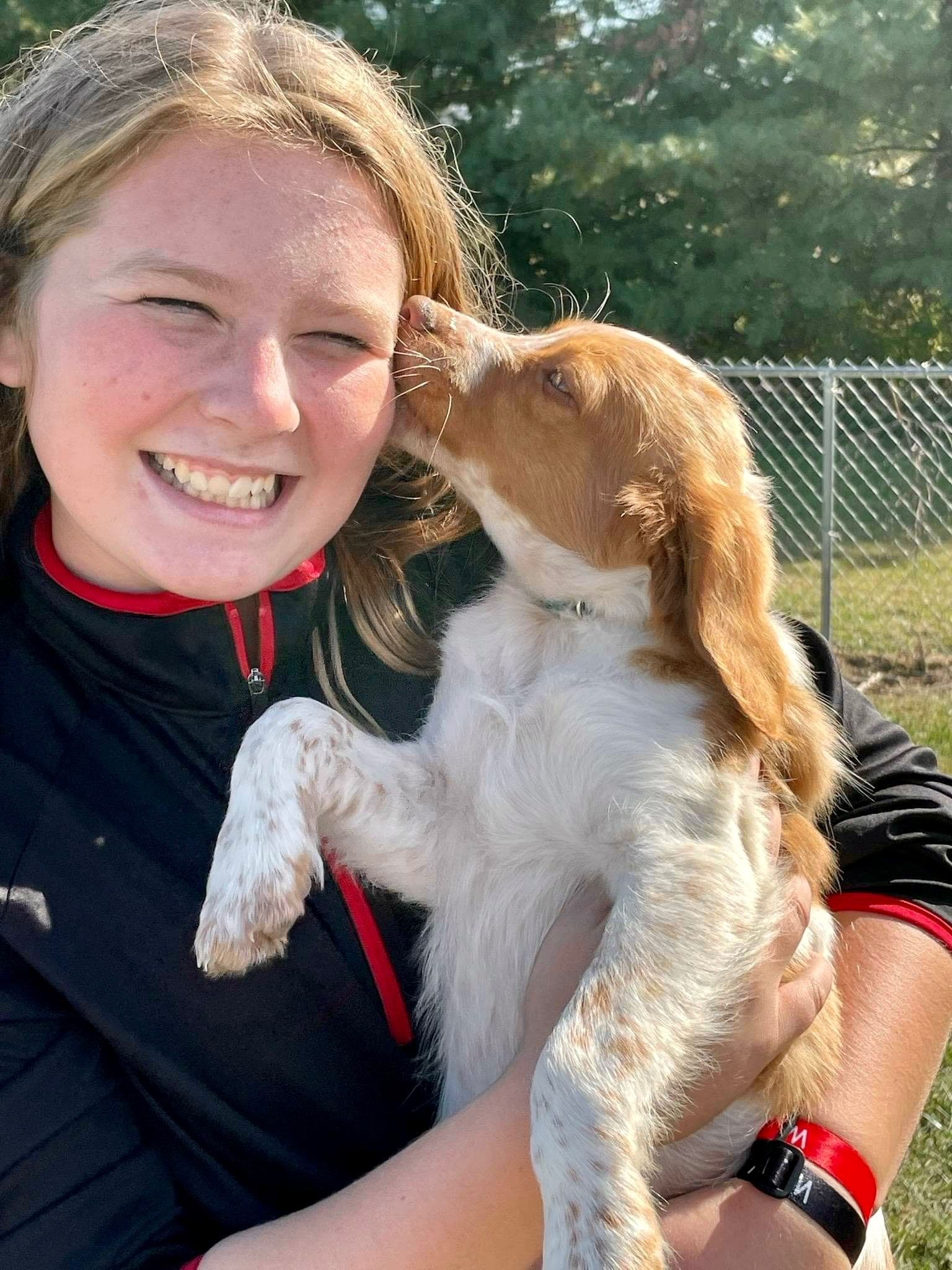 Woman smiling, holding a puppy, getting kissed. Outdoors; grassy area, light hair, black and red clothing.