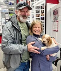 A couple holding a puppy indoors; the woman smiles while embracing the dog.