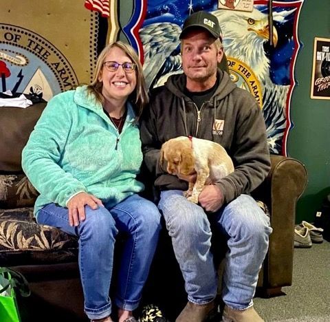 Couple with a small dog sits on a couch indoors. Man holds dog, woman smiles. US Army banner in background.