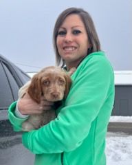 Woman in green jacket holding a puppy, both looking at the camera.