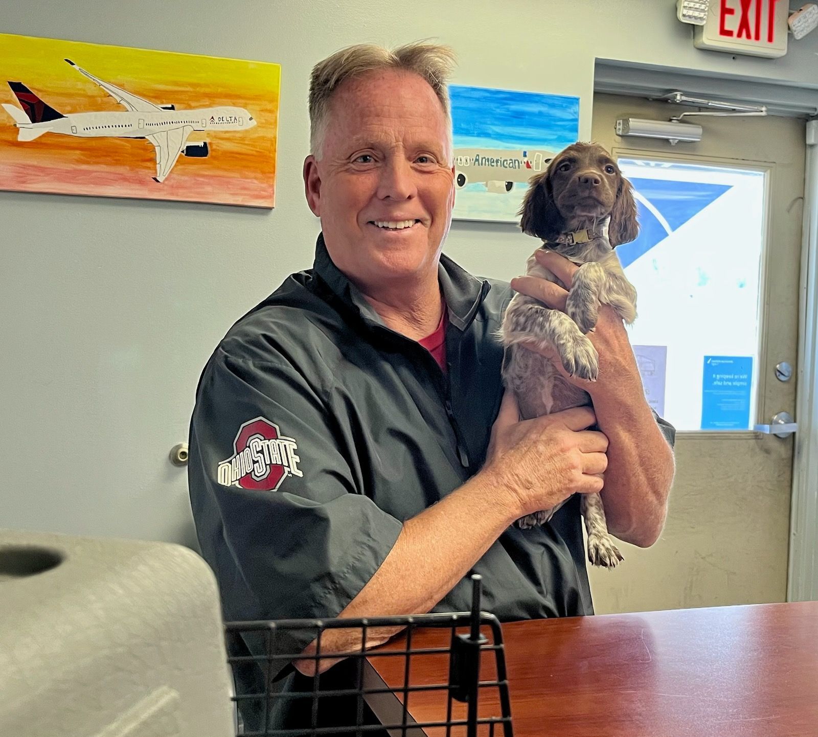 Man holding a small, spotted dog. Behind them are paintings of airplanes and an office setting.